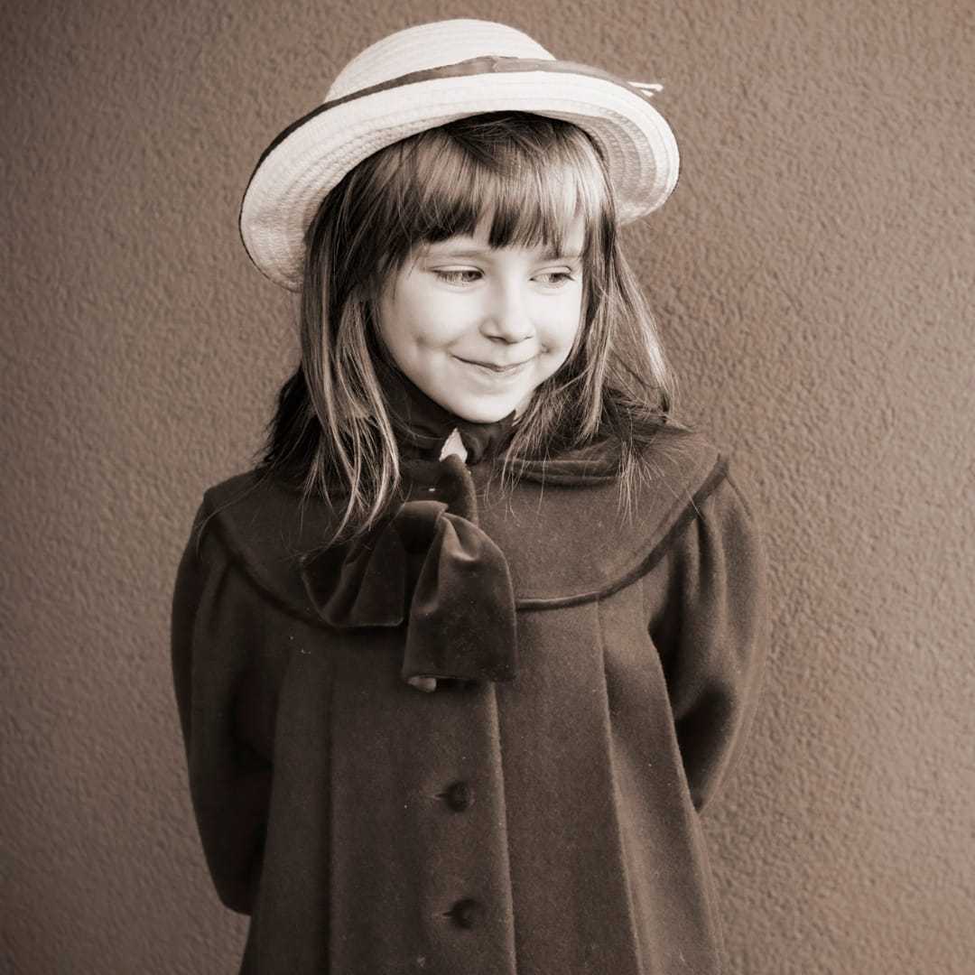 Girl in vintage dress and hat, smiling with hands behind her back. Sepia-toned photo.
