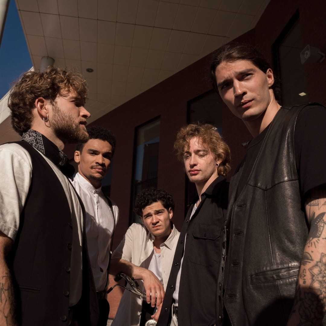 Five men in stylish outfits stand confidently in an urban setting.